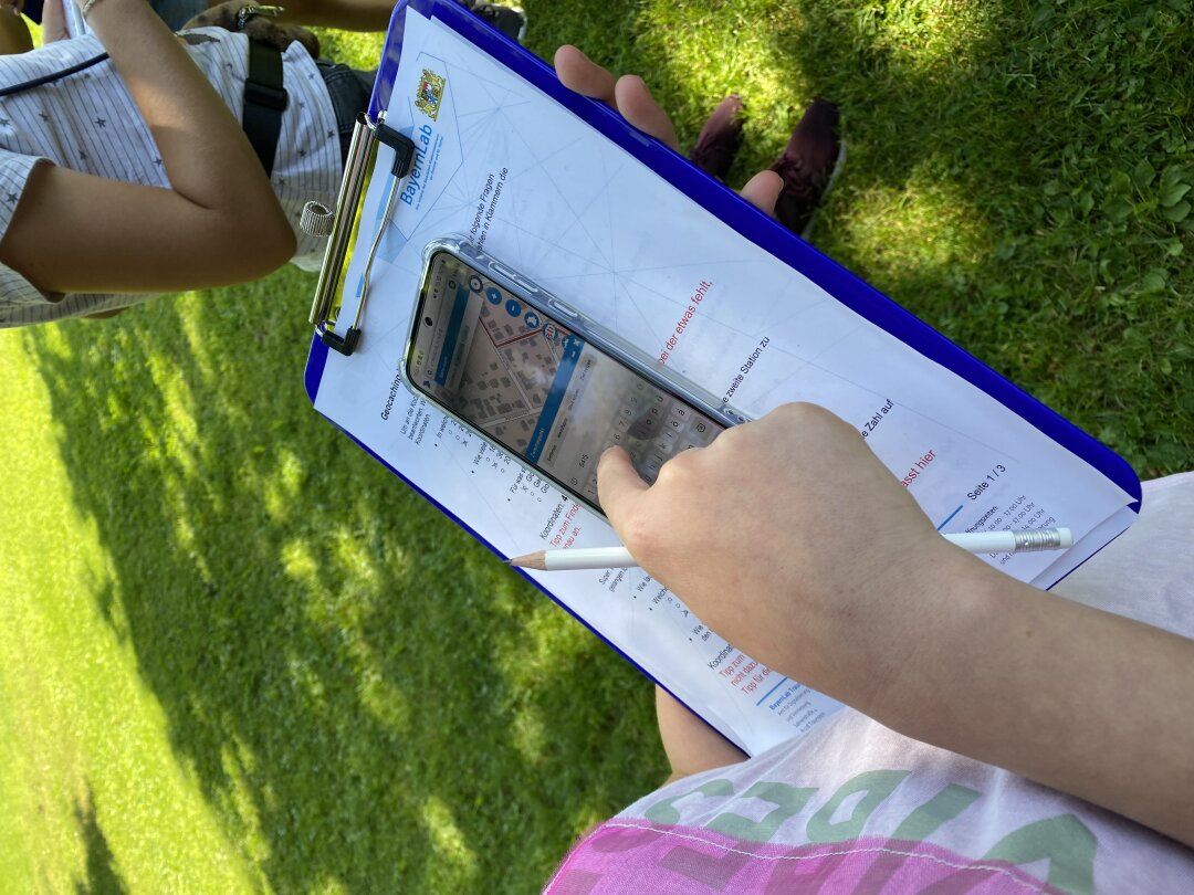 Eine Kinderhand mit einem Bleistift schaut auf ein Clipboard, auf dem ein Blatt Papier eingespannt ist und ein Mobiltelefon mit einem Kartenausschnitt zu sehen ist. Es steht auf einer Wiese.