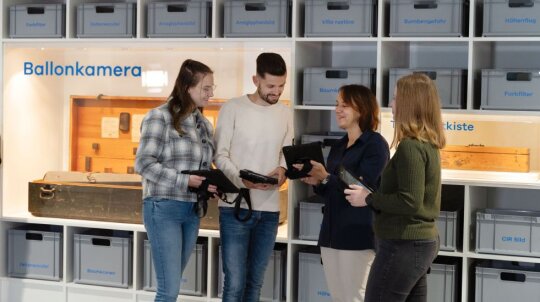 vier Personen vor Regalwand mit grauen Boxen